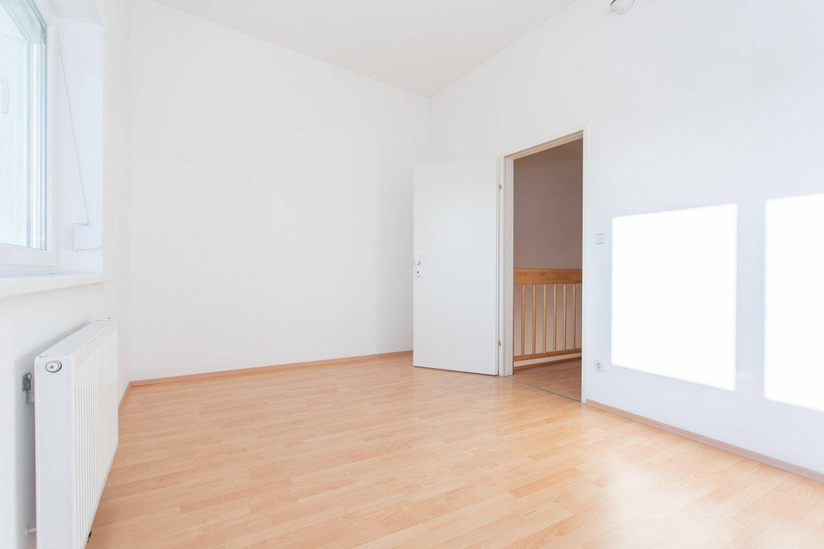 Leerer Raum mit Laminatboden und weiß gestrichenen Wänden, mit offener Tür zu einem Treppenhaus.