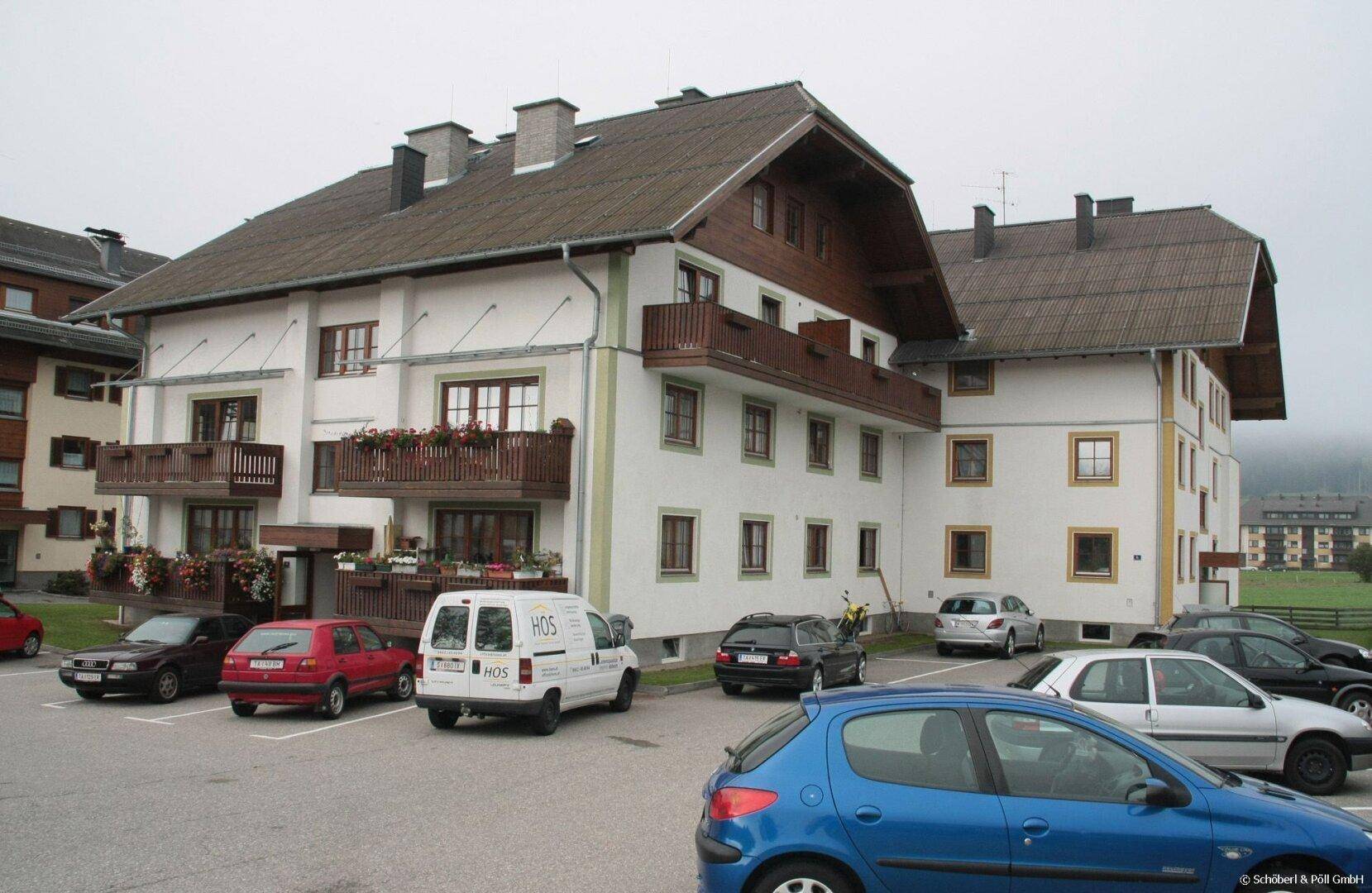 Mehrfamilienhaus mit traditioneller Architektur, Balkonen und Parkplätzen an einem bewölkten Tag.