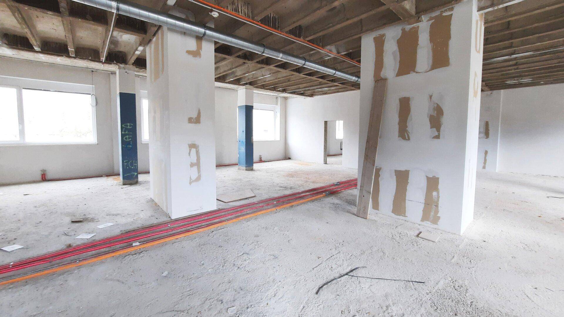 Innenraum im Rohbau mit sichtbaren Deckenbalken, unfertigen Wänden und Installationsleitungen am Boden.