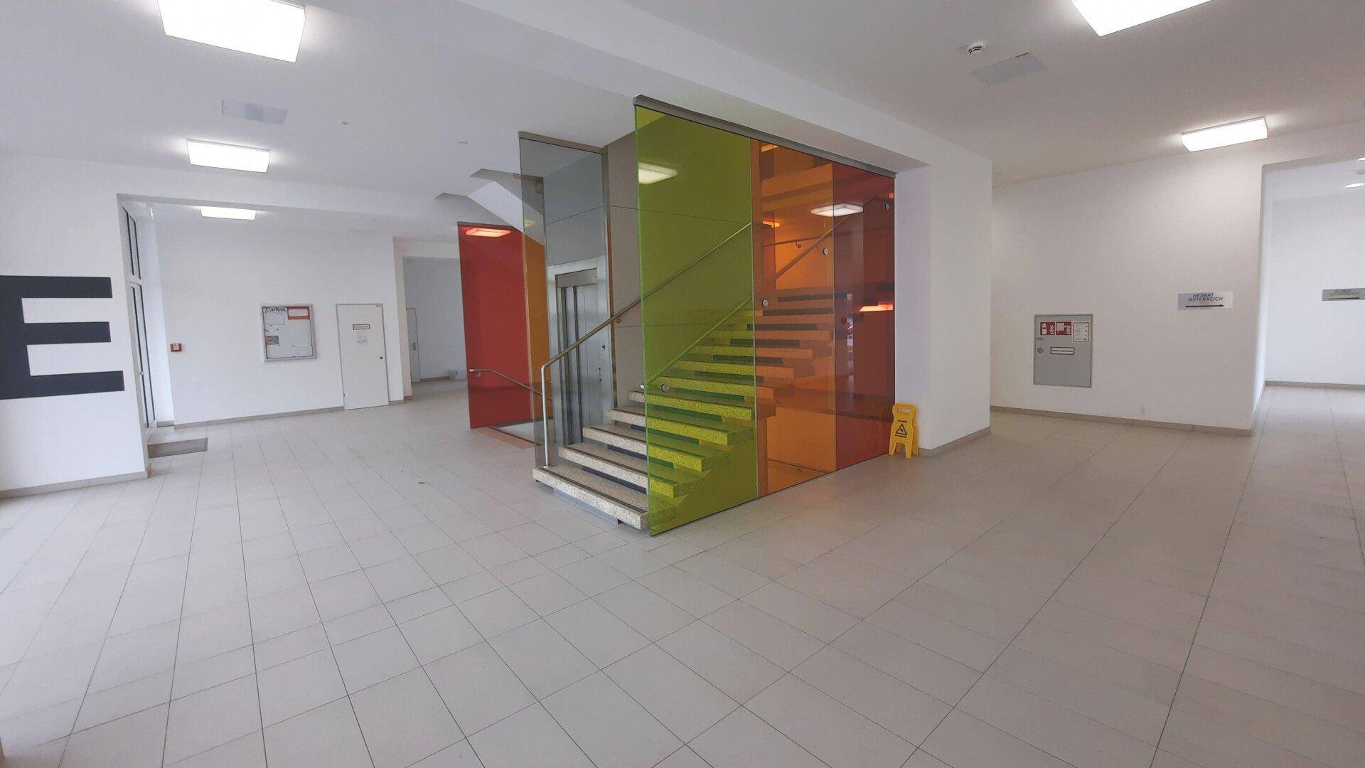 Geräumiger Eingangsbereich mit moderner Treppe, farbigen Glaswänden und einem Aufzug.