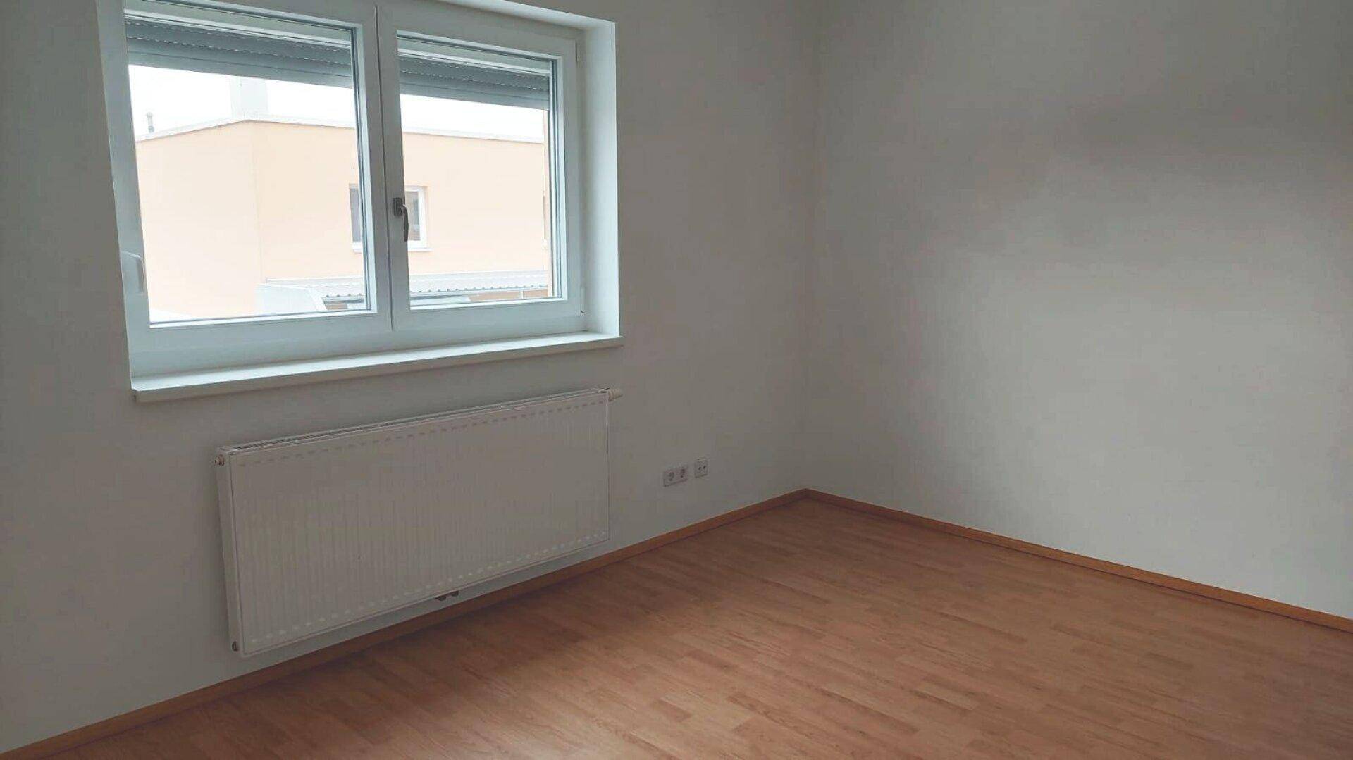 Helles Schlafzimmer mit Laminatboden und Fenster, das einen Blick auf die umliegenden Gebäude freigibt.