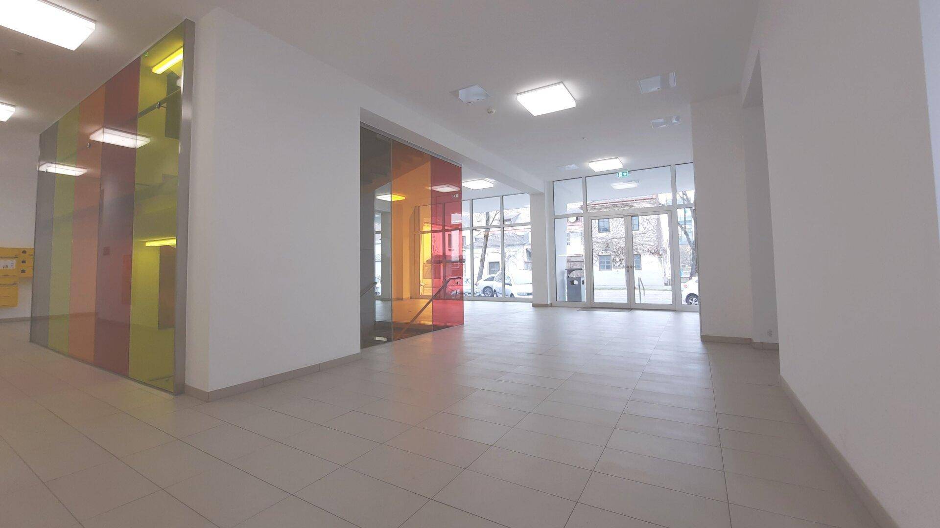 Heller Eingangsbereich mit großen Glasfronten, Fliesenboden und einer auffälligen farbigen Treppenverkleidung.