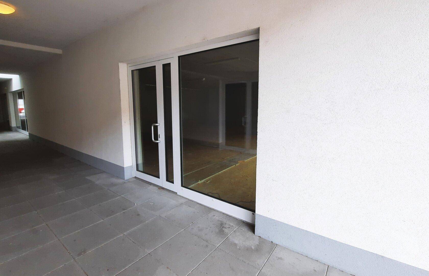 Eingangsbereich mit großer Glastür und Fliesenboden in einem überdachten Gang.