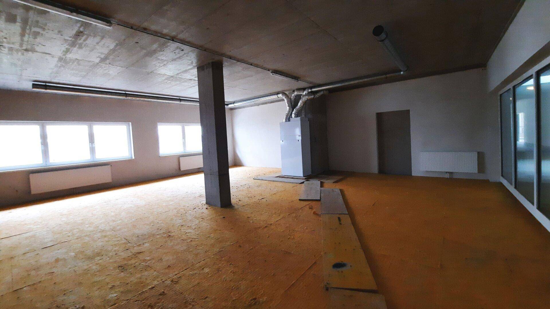 Großer, leerer Raum mit sichtbaren Lüftungsrohren, Betonstütze und großen Fenstern.