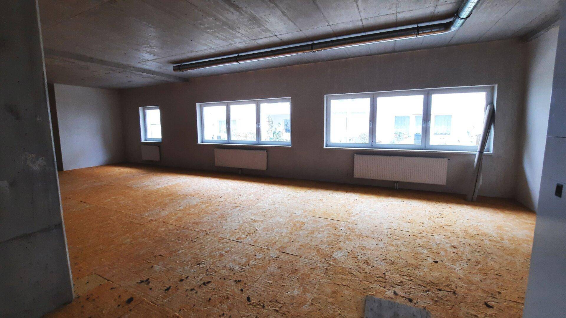 Heller, leerer Raum mit großen Fenstern, die viel Tageslicht hereinlassen, und sichtbaren Rohren.