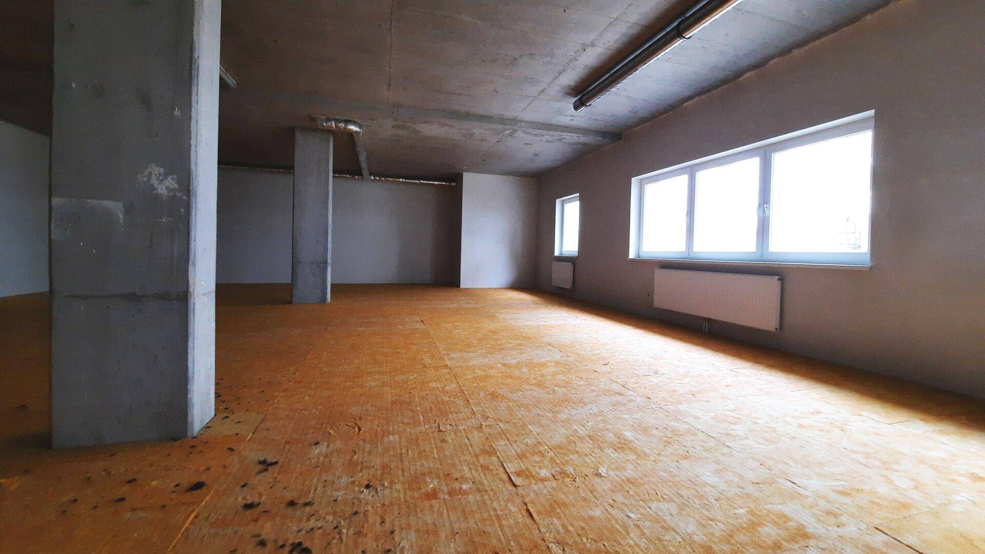 Großer, leerer Raum im Rohbau mit Betonwänden, Fenstern, Heizkörpern und OSB-Unterboden.