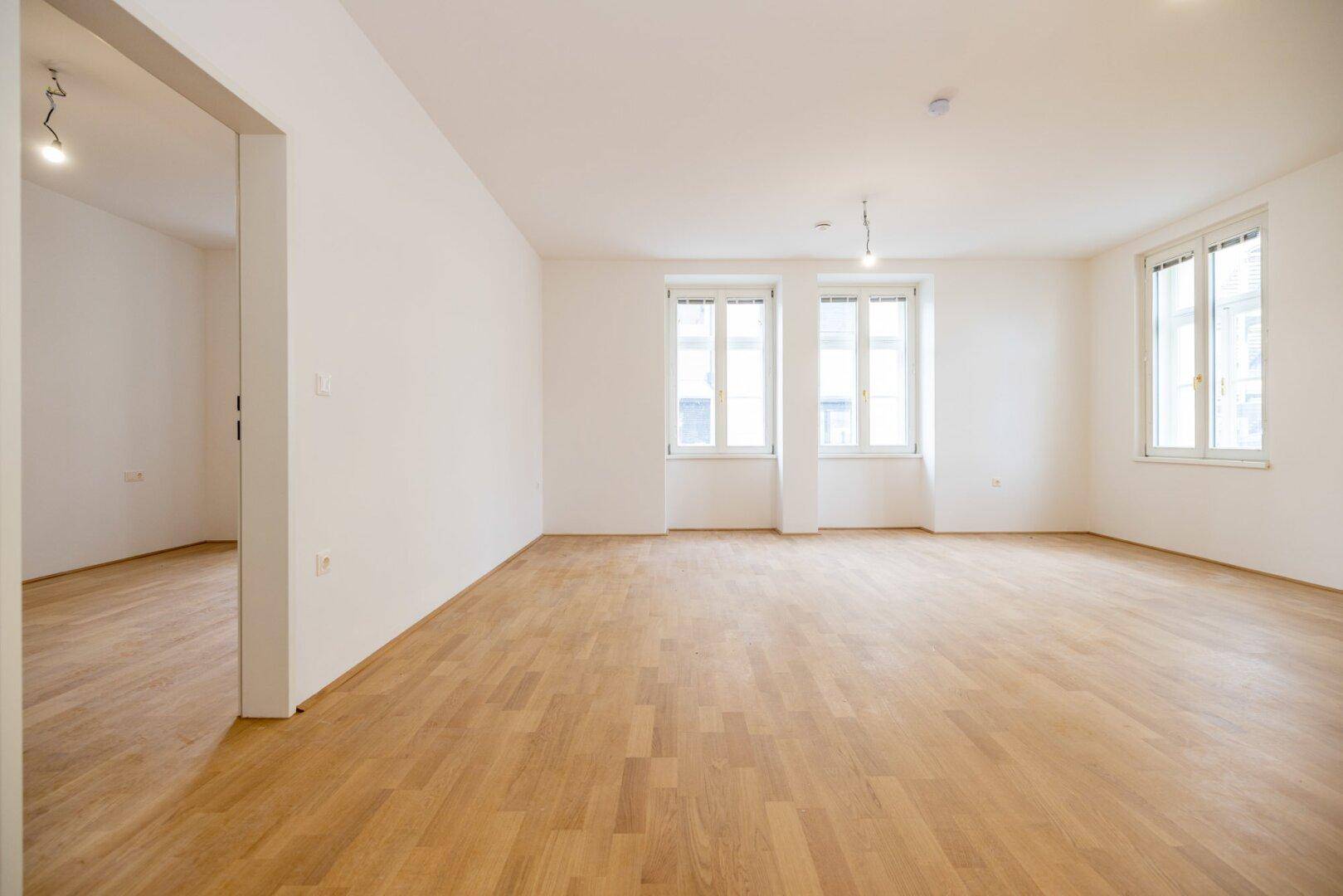 Lichtdurchfluteter Raum mit hellem Parkettboden und großen Fenstern, ideal für einen Wohnbereich.