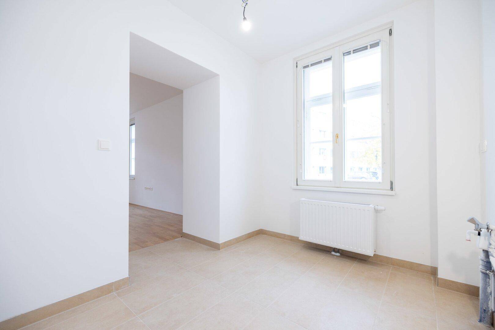 Heller Raum mit Fliesenboden, Fenster und Heizkörper, vorbereitet für eine Küchenzeile.
