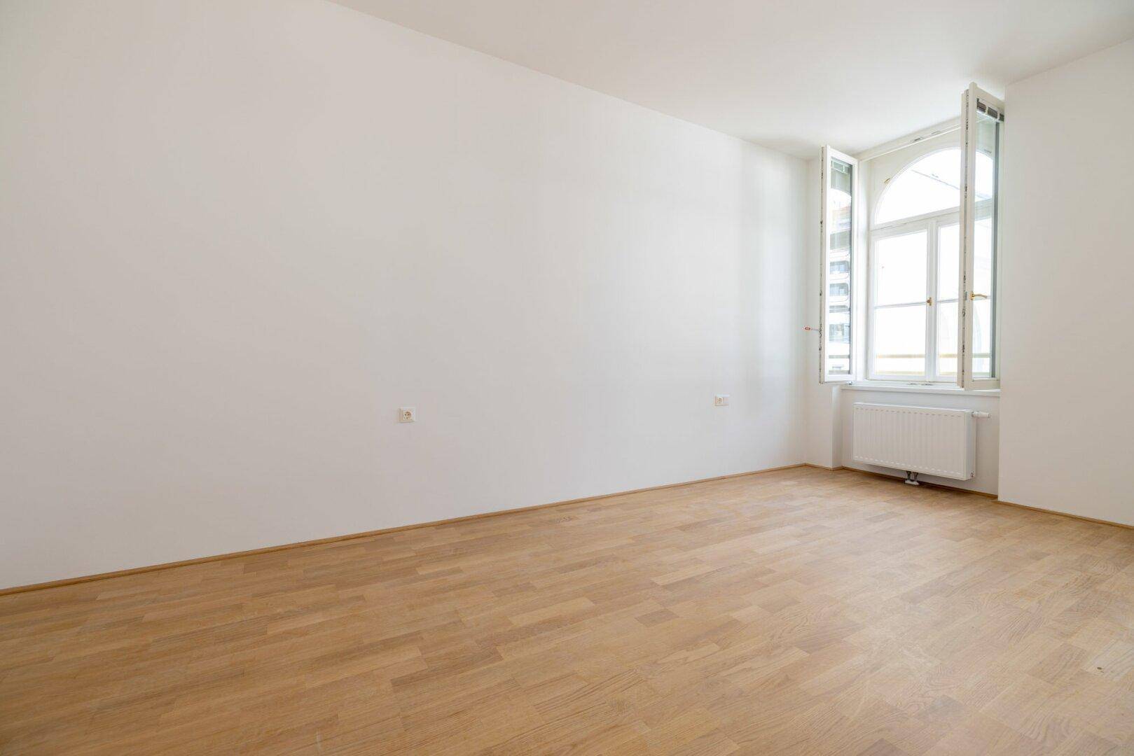 Geräumiges, helles Zimmer mit Holzboden und einem großen Fenster, ideal für Wohn- oder Schlafbereich.