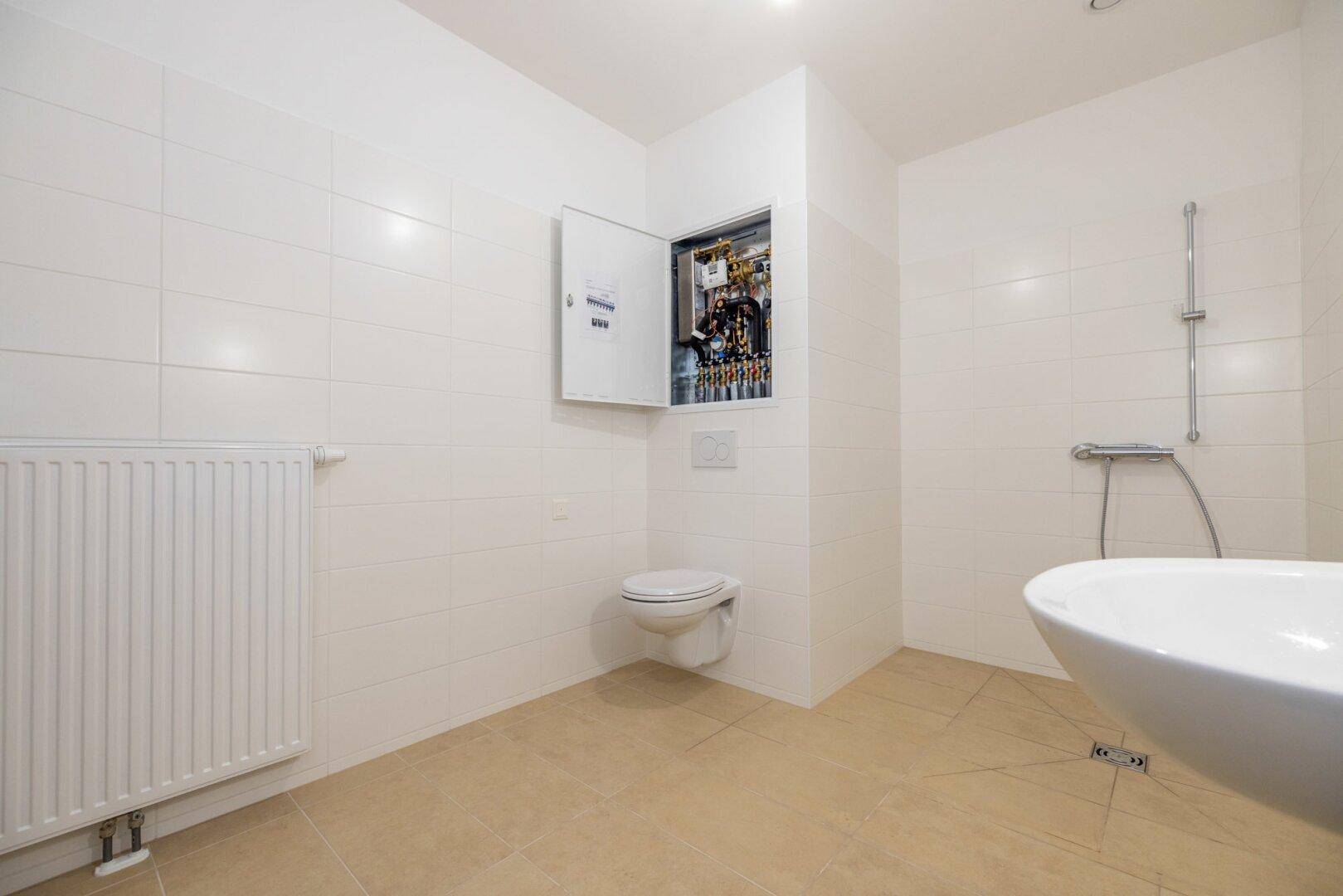 Helles Badezimmer mit weißer Fliesenwand, Heizkörper und Duschbereich, in gepflegtem Zustand.