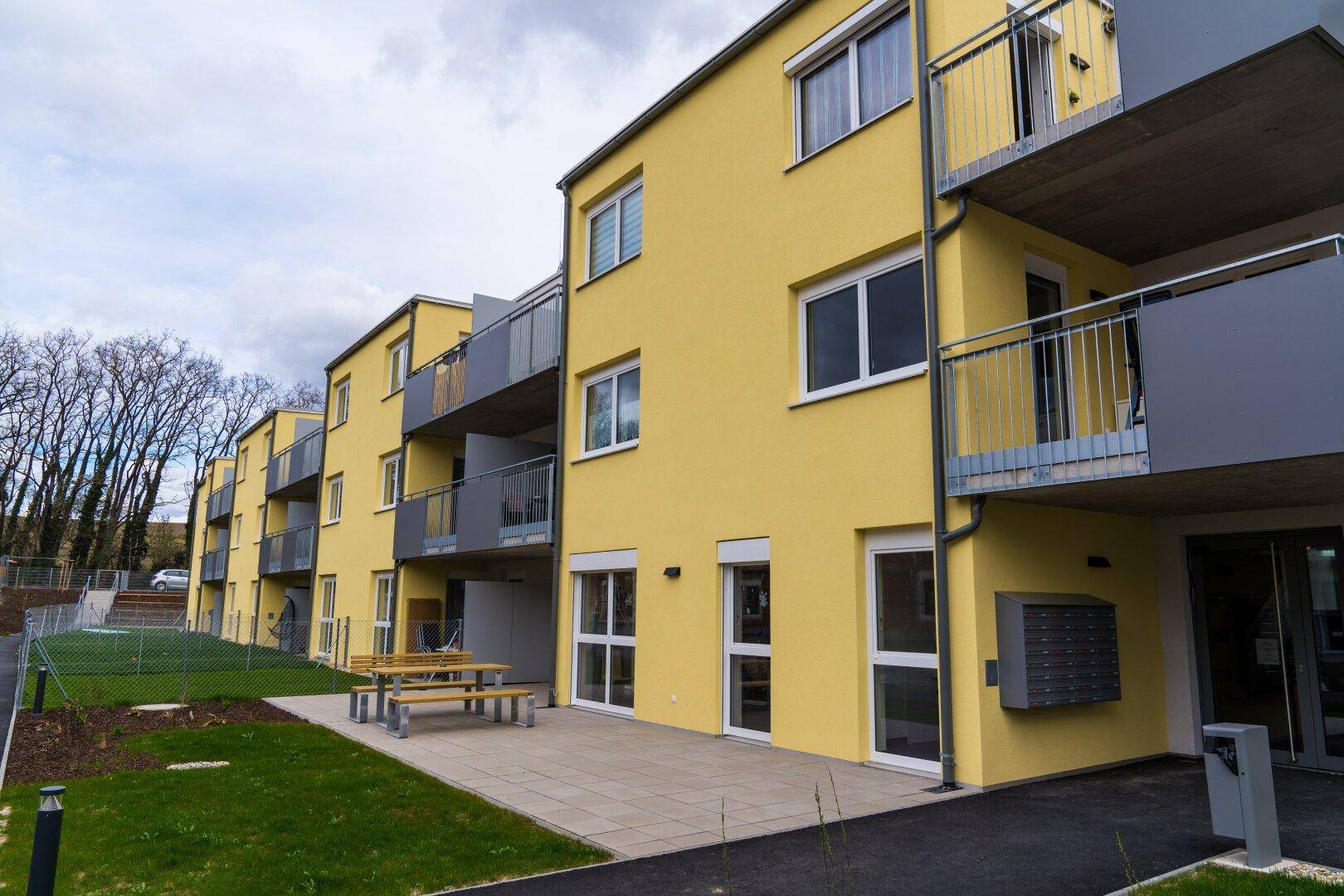 Moderne Wohnanlage mit gelber Fassade, Balkonen und einer gemeinschaftlichen Terrasse mit Sitzgelegenheiten.