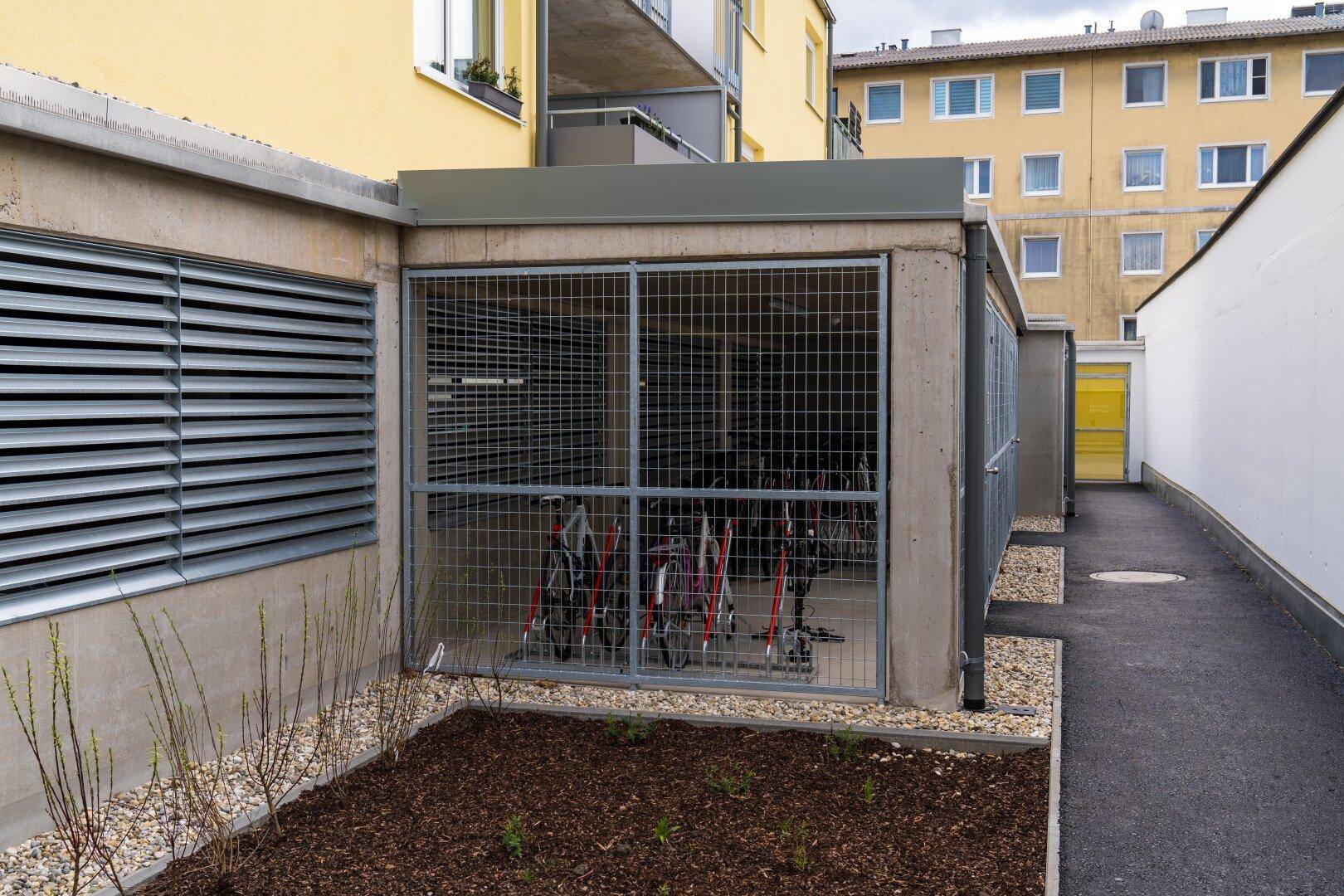 Sicherer Fahrradabstellraum mit Gittertüren und angrenzender Grünfläche im Außenbereich.