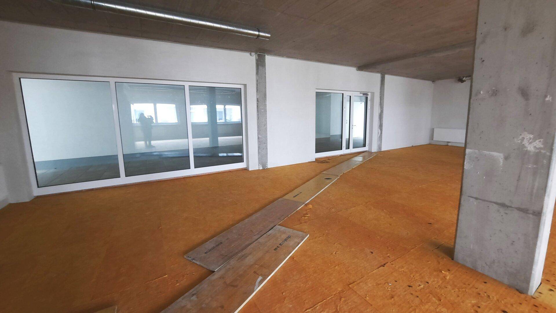 Geräumiger, offener Innenraum mit großen Fenstern, unfertigem Bodenbelag und sichtbaren Betonelementen, ideal für ein Büro.