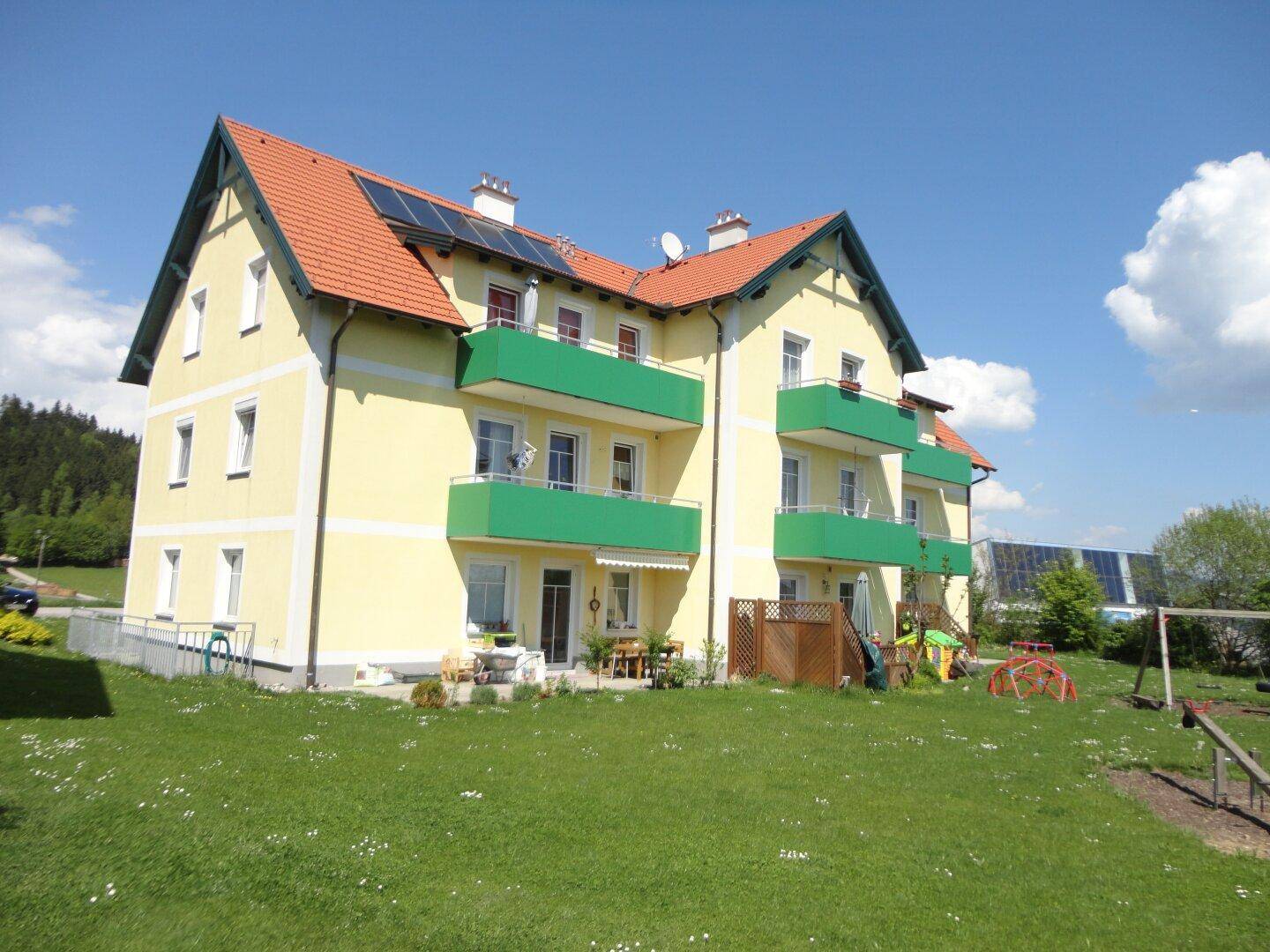 Mehrfamilienhaus mit gelber Fassade, grünen Balkonen und rotem Ziegeldach, umgeben von einer grünen Wiese mit Spielplatz.