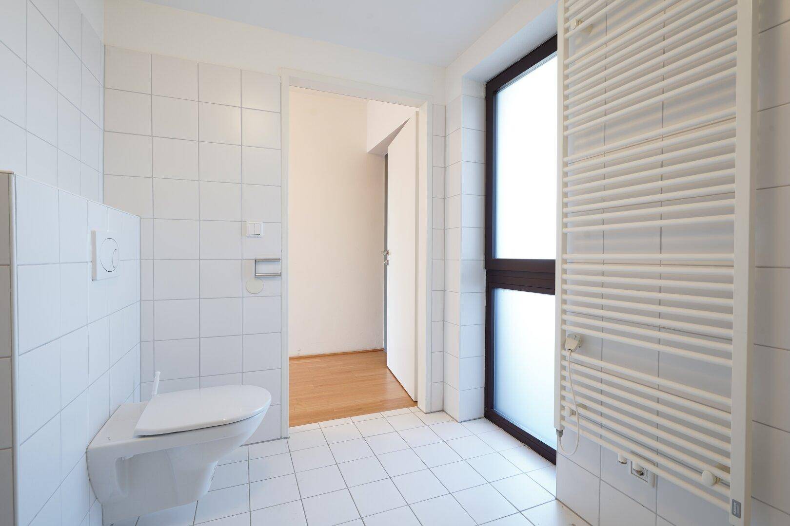 Badezimmer mit wandhängendem WC, Fenster und Heizkörper, der auch als Handtuchhalter dient.