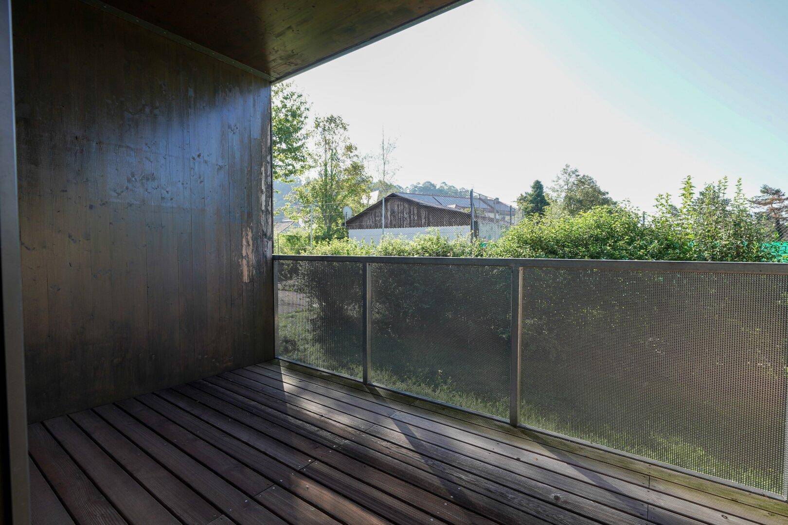 Sonniger Balkon mit Holzboden und Blick ins Grüne, umgeben von Bäumen.