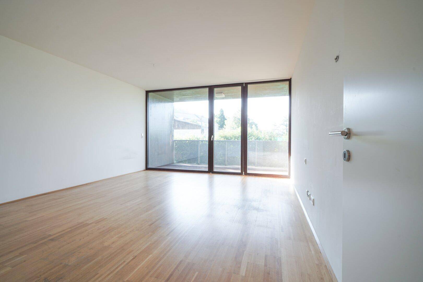 Lichtdurchfluteter Raum mit Holzboden und breiter Fensterfront, die zum Balkon führt.