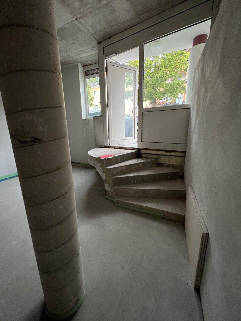 Eingangsbereich im Rohbau mit einer Wendeltreppe und Blick nach draußen durch die Tür.