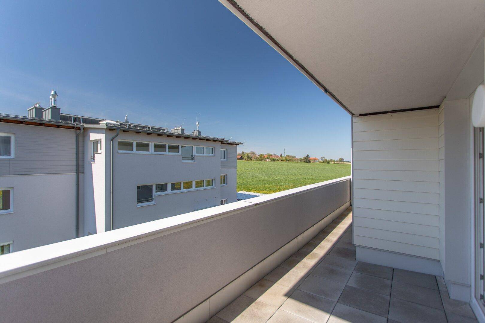 Langer Balkon mit Fliesenboden und Blick auf ein angrenzendes Feld und weitere Gebäude.