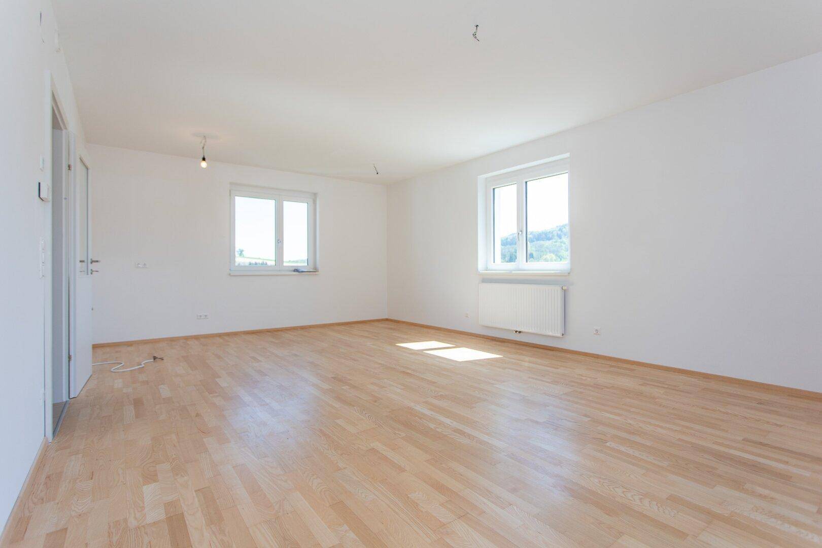 Großes, leeres Zimmer mit hellem Parkettboden und zwei Fenstern, die viel Licht hereinlassen.