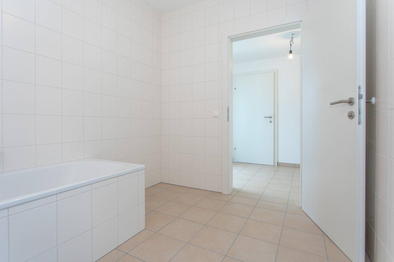 Helles Badezimmer mit weißen Wandfliesen und beigem Boden, bietet eine Badewanne.