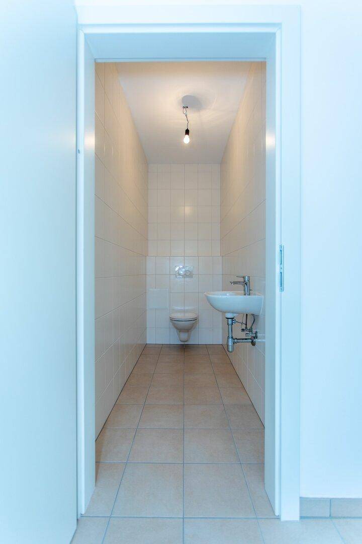 Schmale Gästetoilette mit Wand-WC, kleinem Waschbecken und hellen Fliesen an Wänden und Boden.