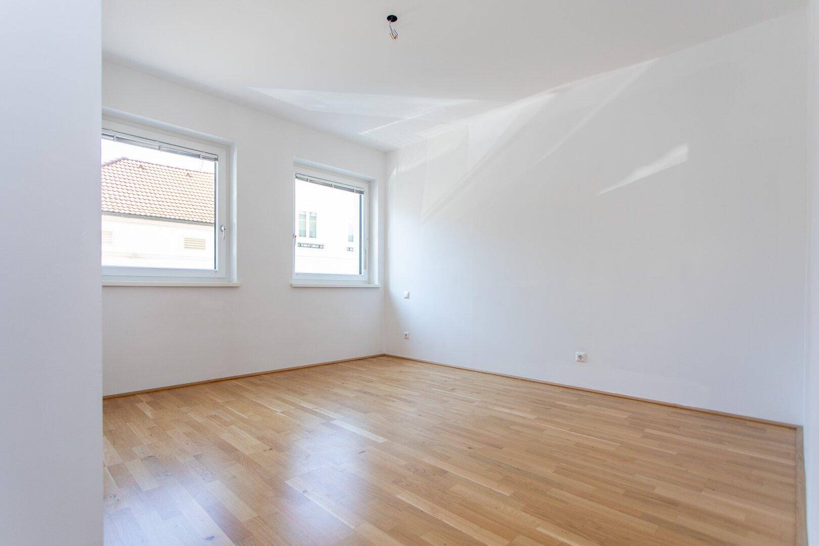 Helles Schlafzimmer mit zwei Fenstern und glänzendem Parkettboden, bereit zur Einrichtung.