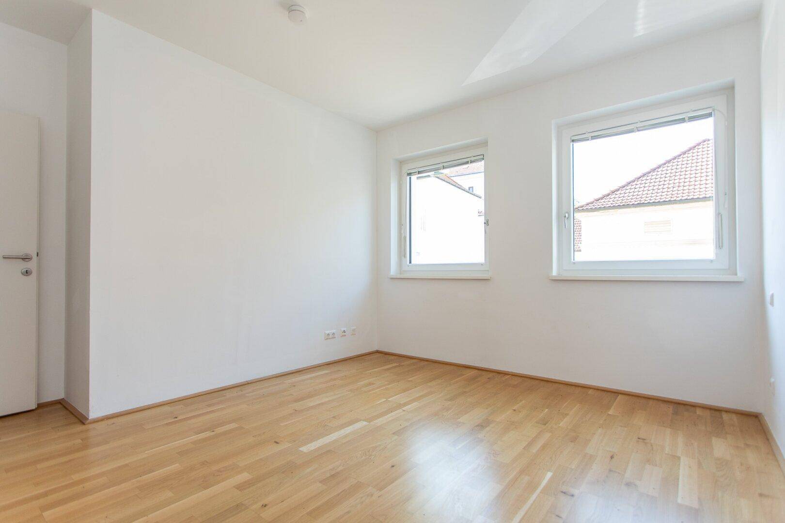 Unmöbliertes Schlafzimmer mit Parkettboden und zwei Fenstern, die viel Tageslicht hereinlassen.