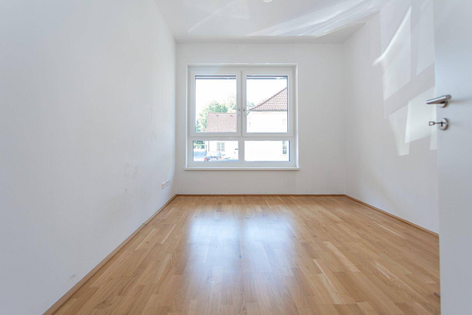 Freundliches Schlafzimmer mit großem Fenster und hellem Parkettboden, ideal für eine ruhige Atmosphäre.