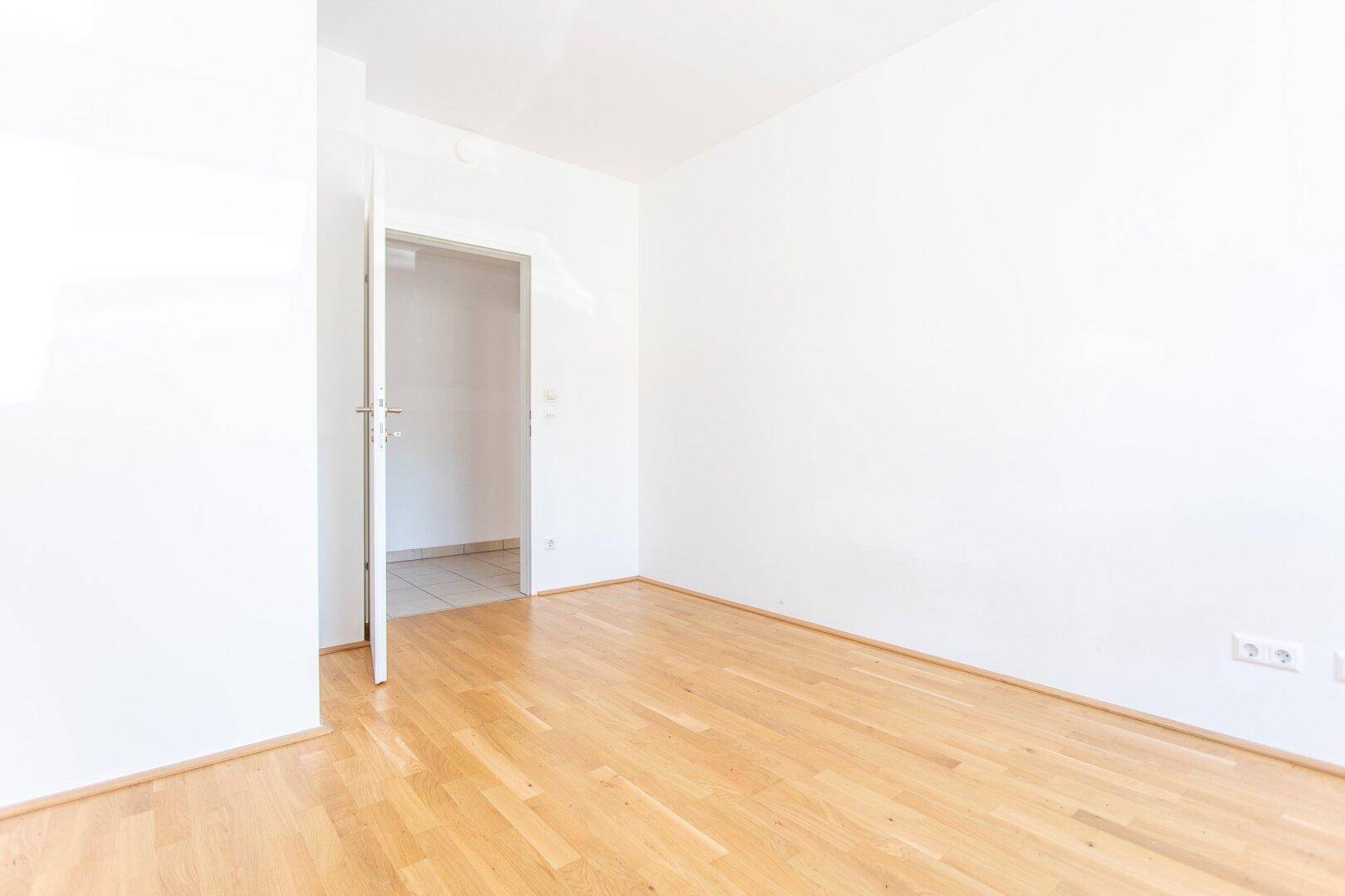 Leeres Zimmer mit Parkettboden und weiß gestrichenen Wänden, durch eine Tür zugänglich.