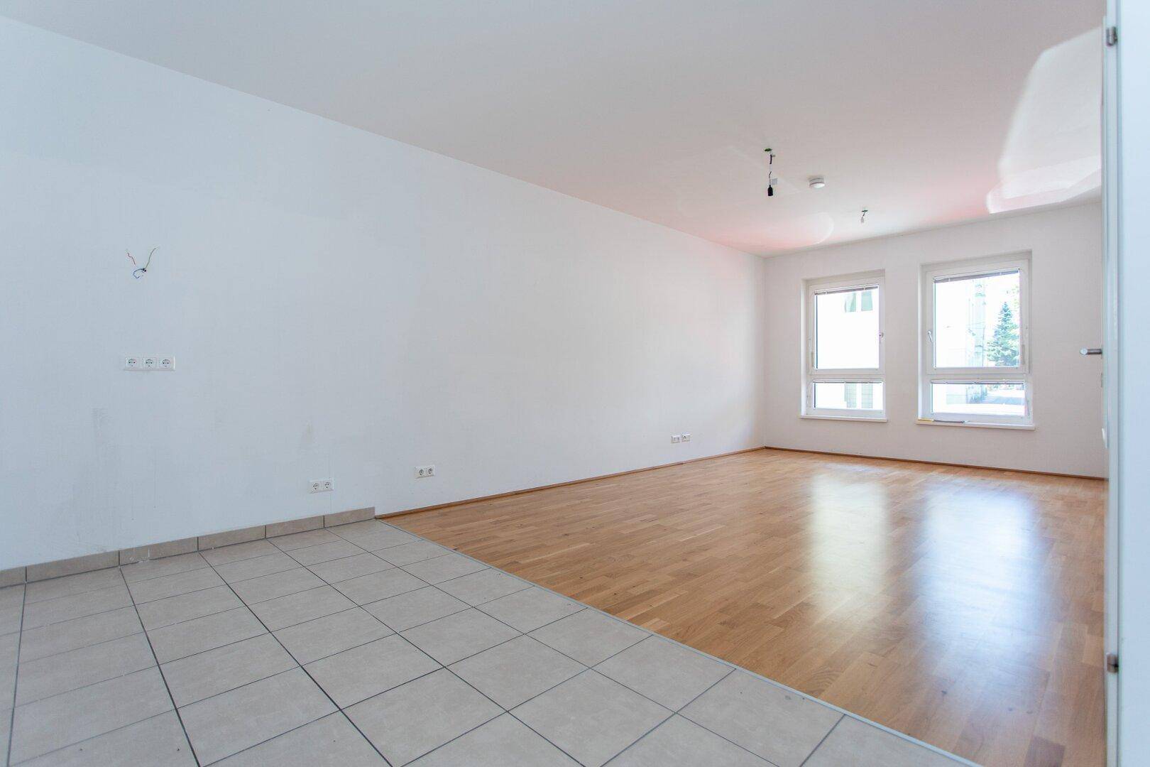Geräumiges, unmöbliertes Wohnzimmer mit großen Fenstern, Parkettboden und einem gefliesten Bereich.