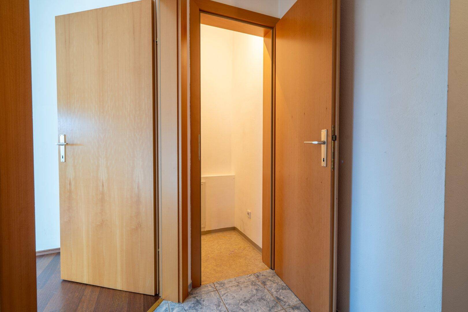 Flur mit Fliesenboden und zwei Holztüren, eine davon führt zu einem kleinen Abstellraum.