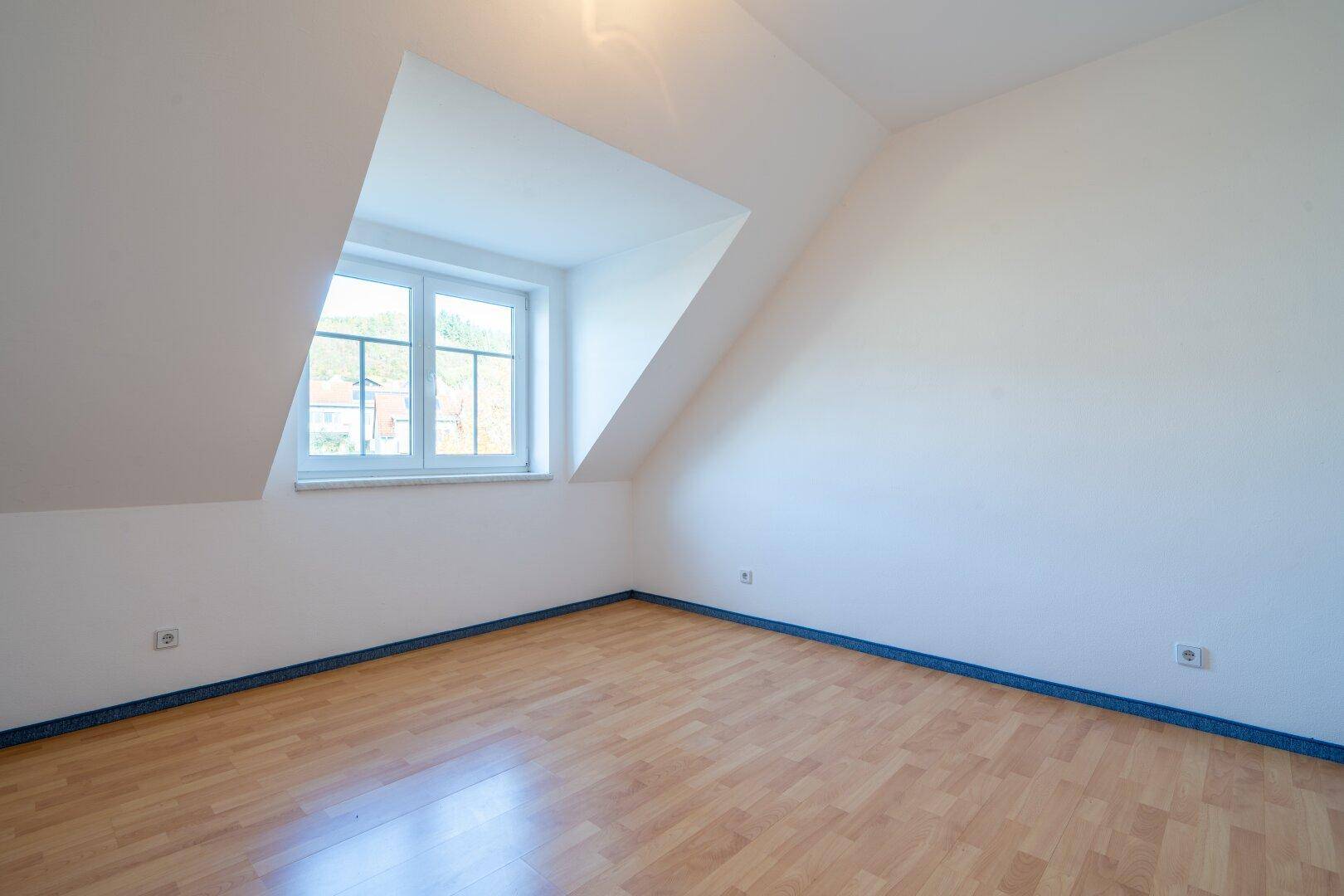 Leeres Zimmer mit hellem Laminatboden, Dachschräge und einem Fenster, das Tageslicht spendet.