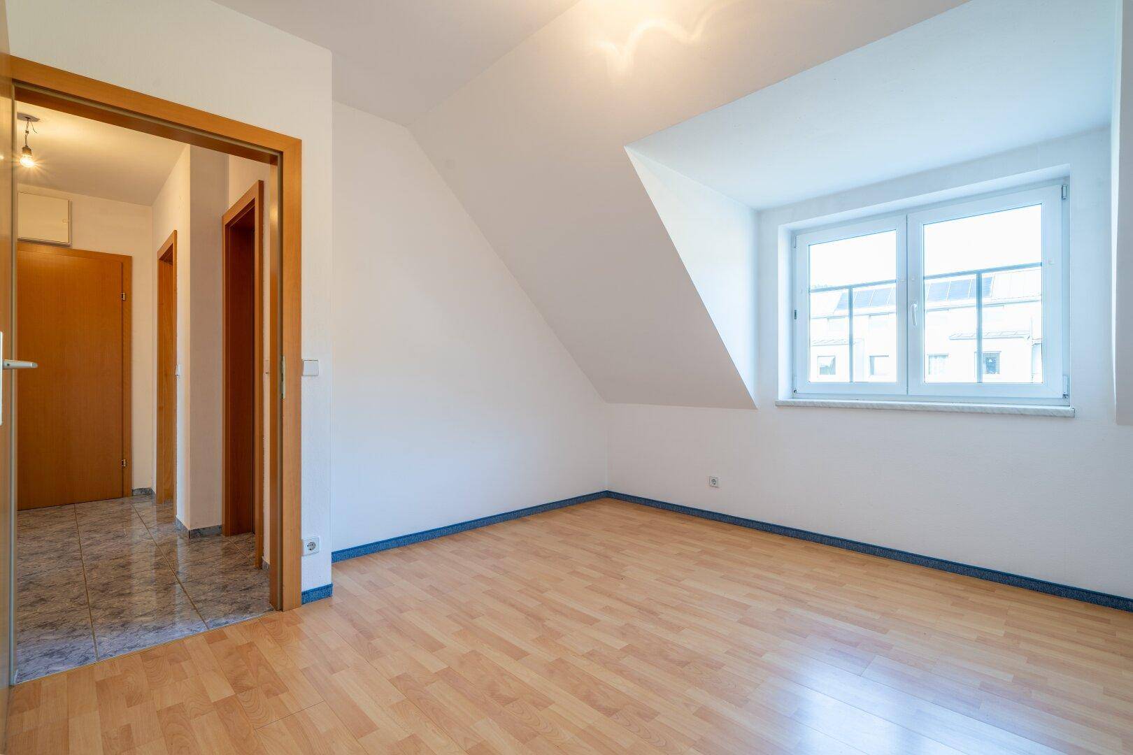 Leeres Zimmer mit hellem Laminatboden, Dachschräge und einer Holztür, die in den Flur führt.
