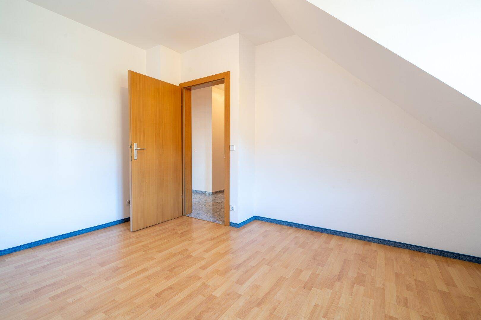 Leeres Zimmer mit hellem Laminatboden, Dachschräge und einer offenen Holztür.