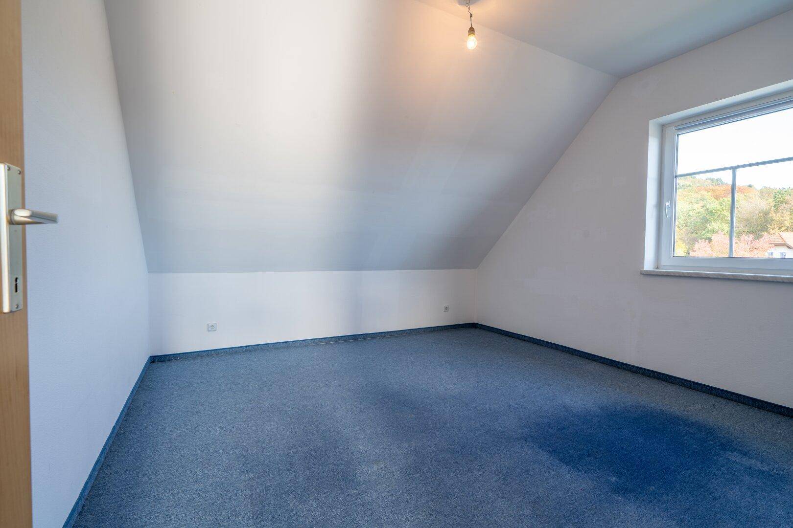 Leeres Zimmer mit blauem Teppichboden, Dachschräge und einem Fenster, das Tageslicht hereinlässt.