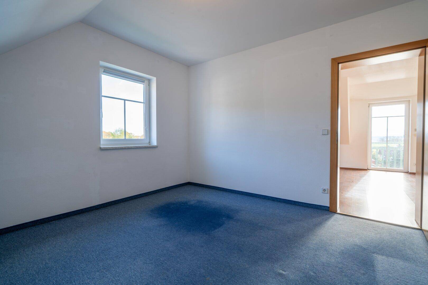 Leeres Zimmer mit blauem Teppichboden, Dachschräge und einem Fenster.