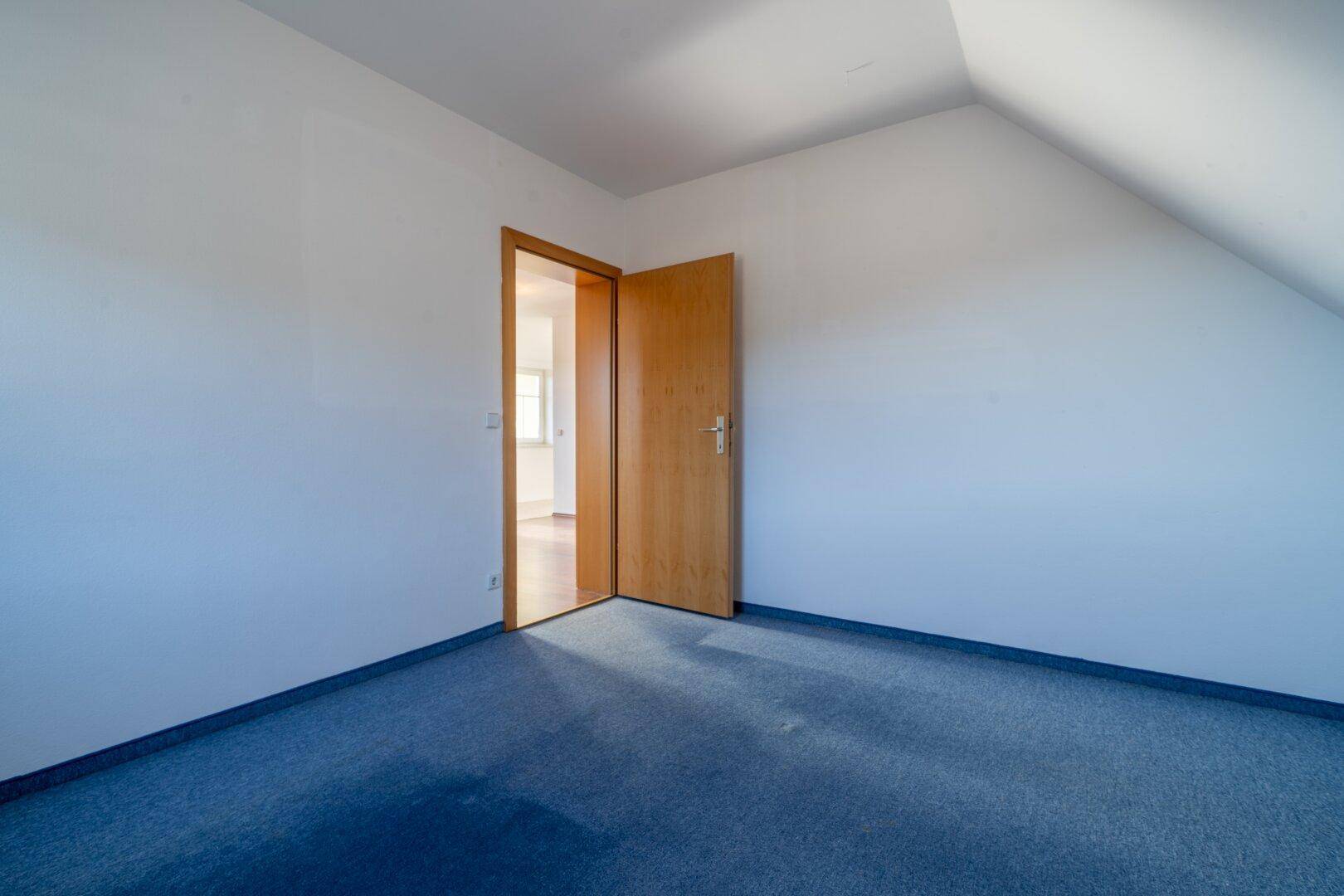 Leeres Zimmer mit blauem Teppichboden, Dachschräge und einer Holztür.