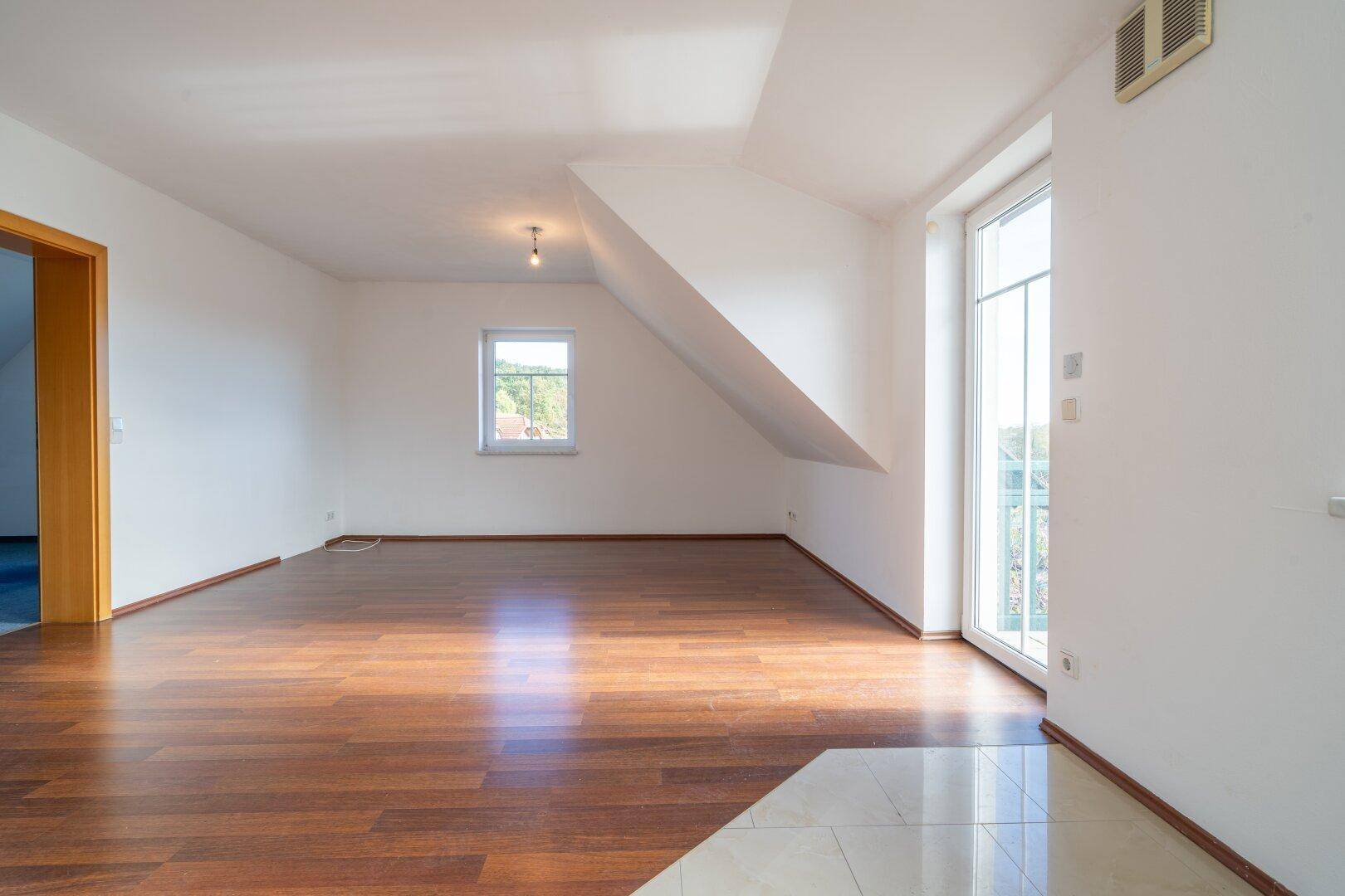 Geräumiger, leerer Raum mit Holzboden, Dachschräge und Balkontür, die viel Licht hereinlässt.