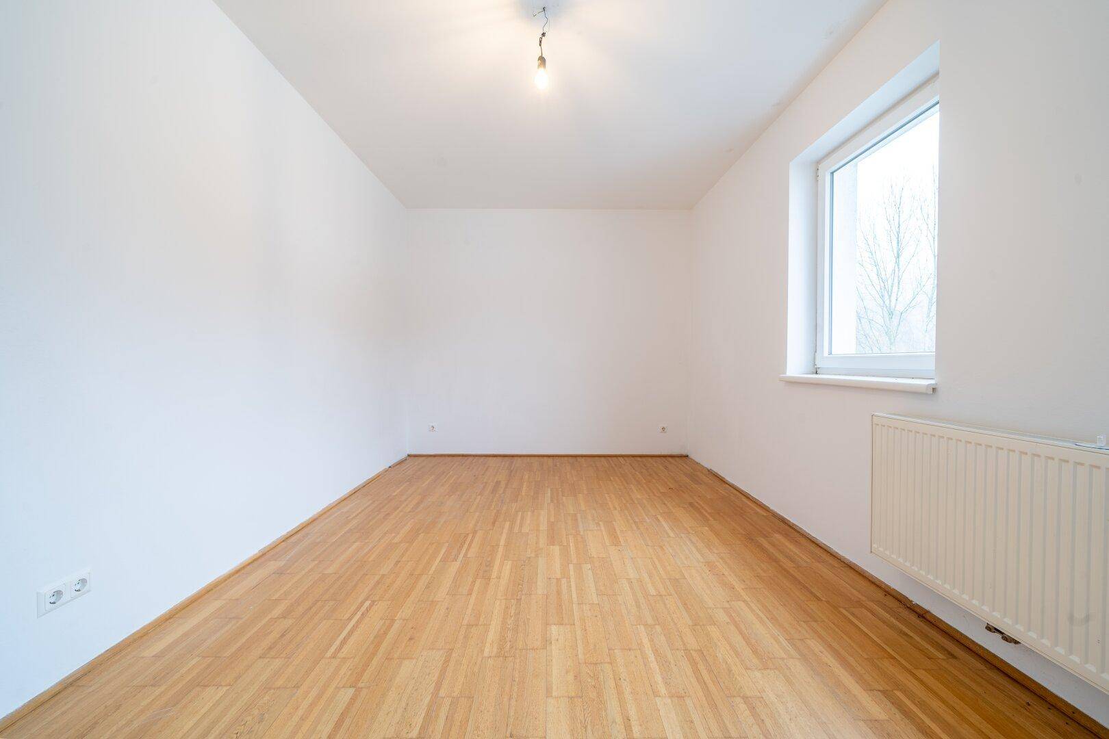Schlichtes Zimmer mit Parkettboden und einem Fenster, bereit zur individuellen Gestaltung.