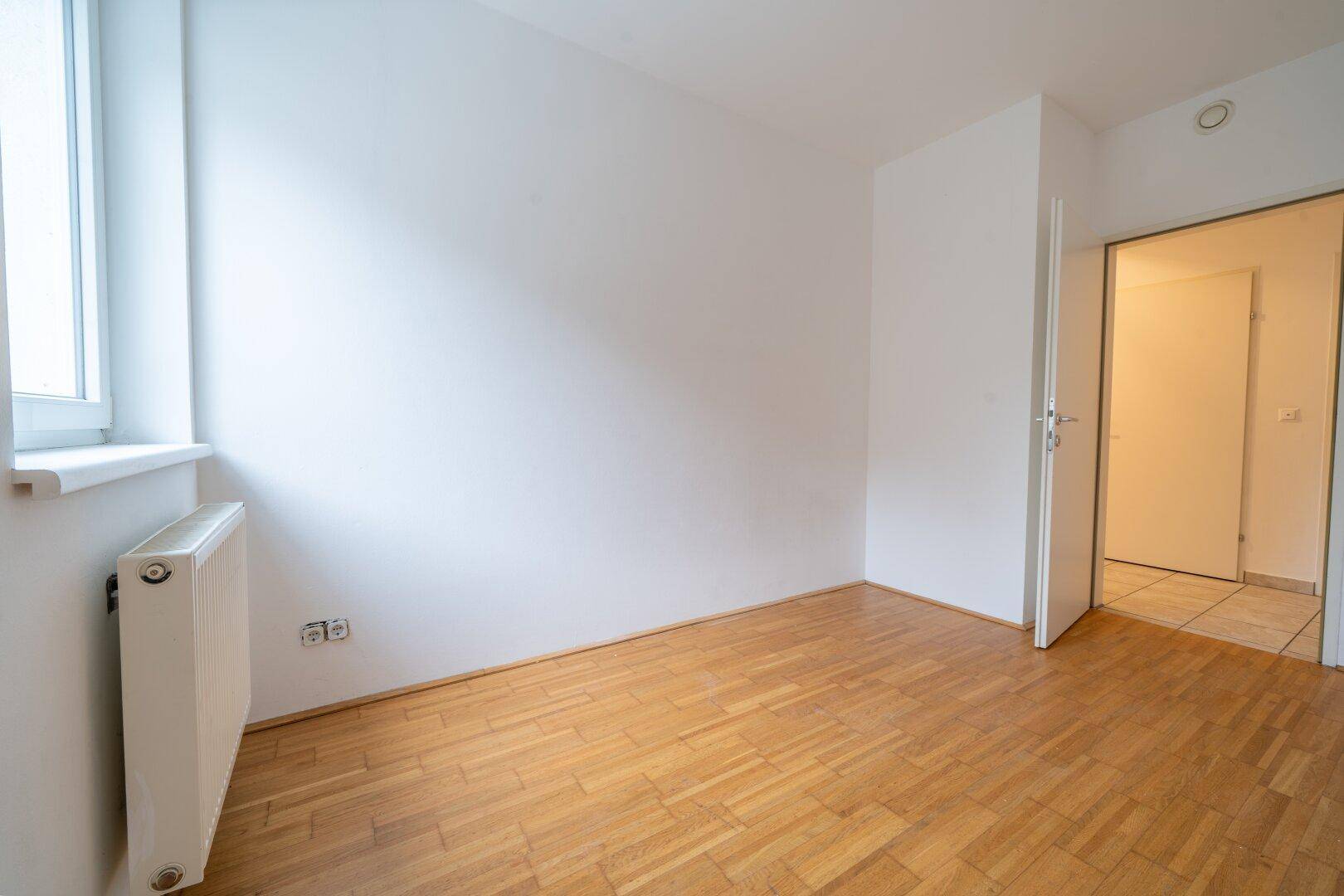 Helles Zimmer mit Parkettboden, einem Fenster und einem Heizkörper, ideal für individuelle Gestaltung.