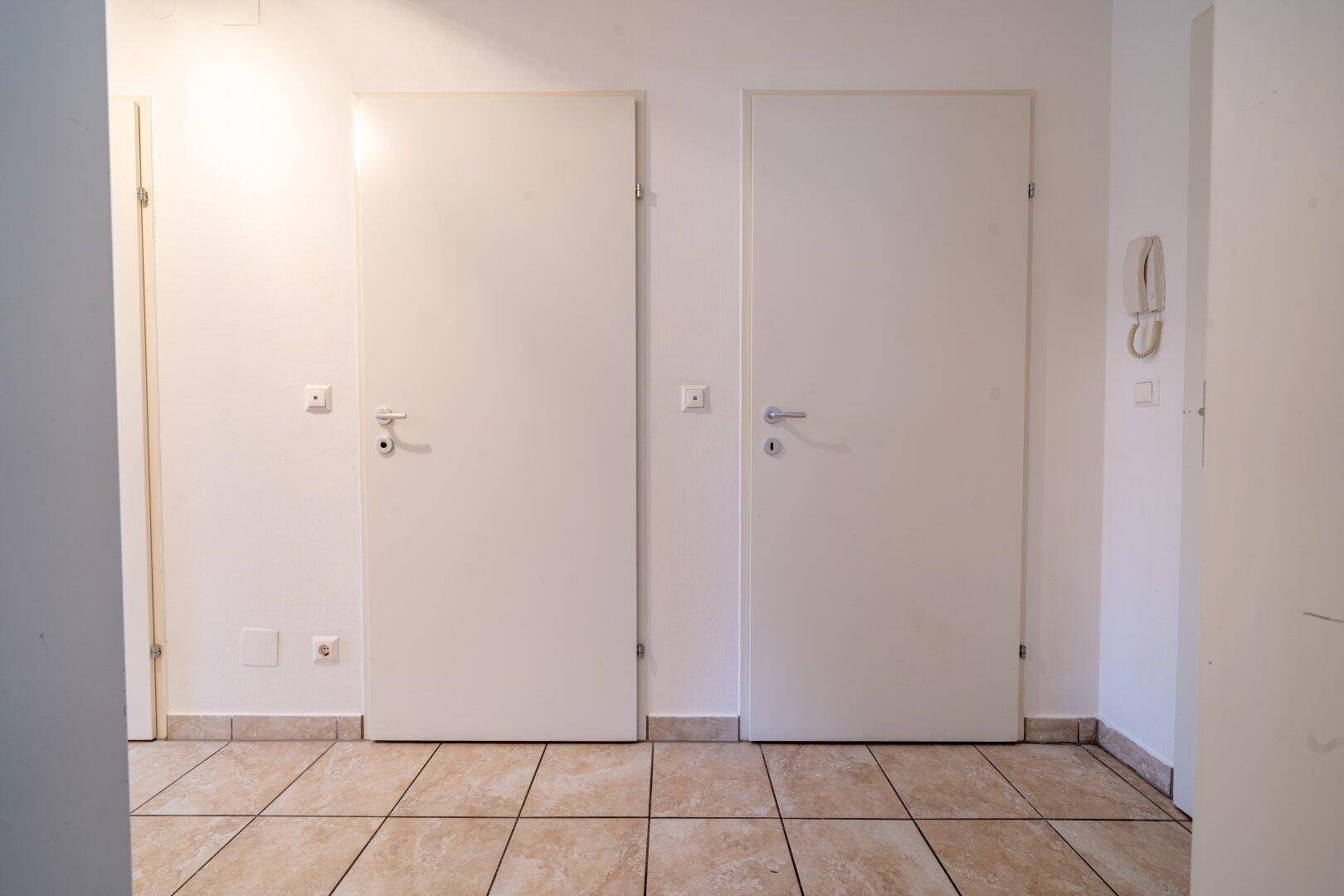 Flur mit zwei weißen Türen und beigefarbenen Fliesen, bietet Zugang zu verschiedenen Bereichen.