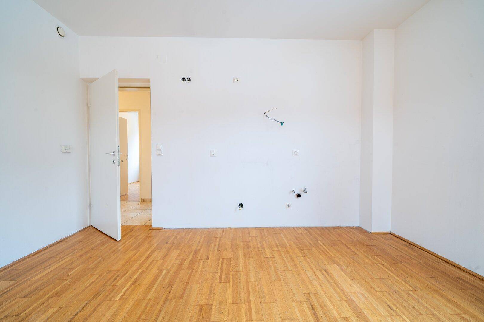 Leeres Zimmer mit Parkettboden und weiß gestrichenen Wänden, mit Tür zu einem weiteren Raum.