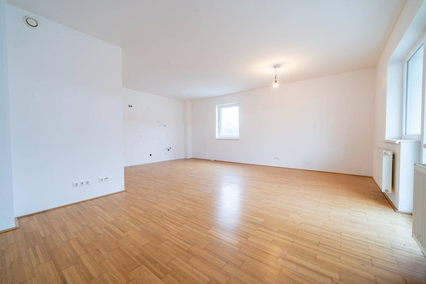 Geräumiges Wohnzimmer mit hellem Parkettboden und mehreren Fenstern für viel Tageslicht.