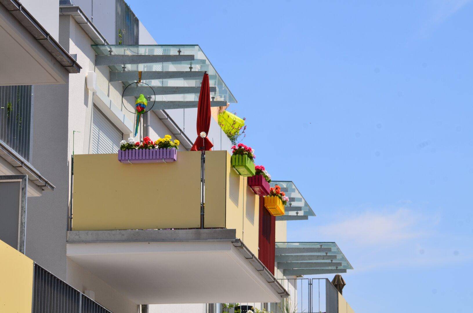 Nahaufnahme von Balkonen mit bunten Blumenkästen und transparenten Glasdächern an einer modernen Fassade.