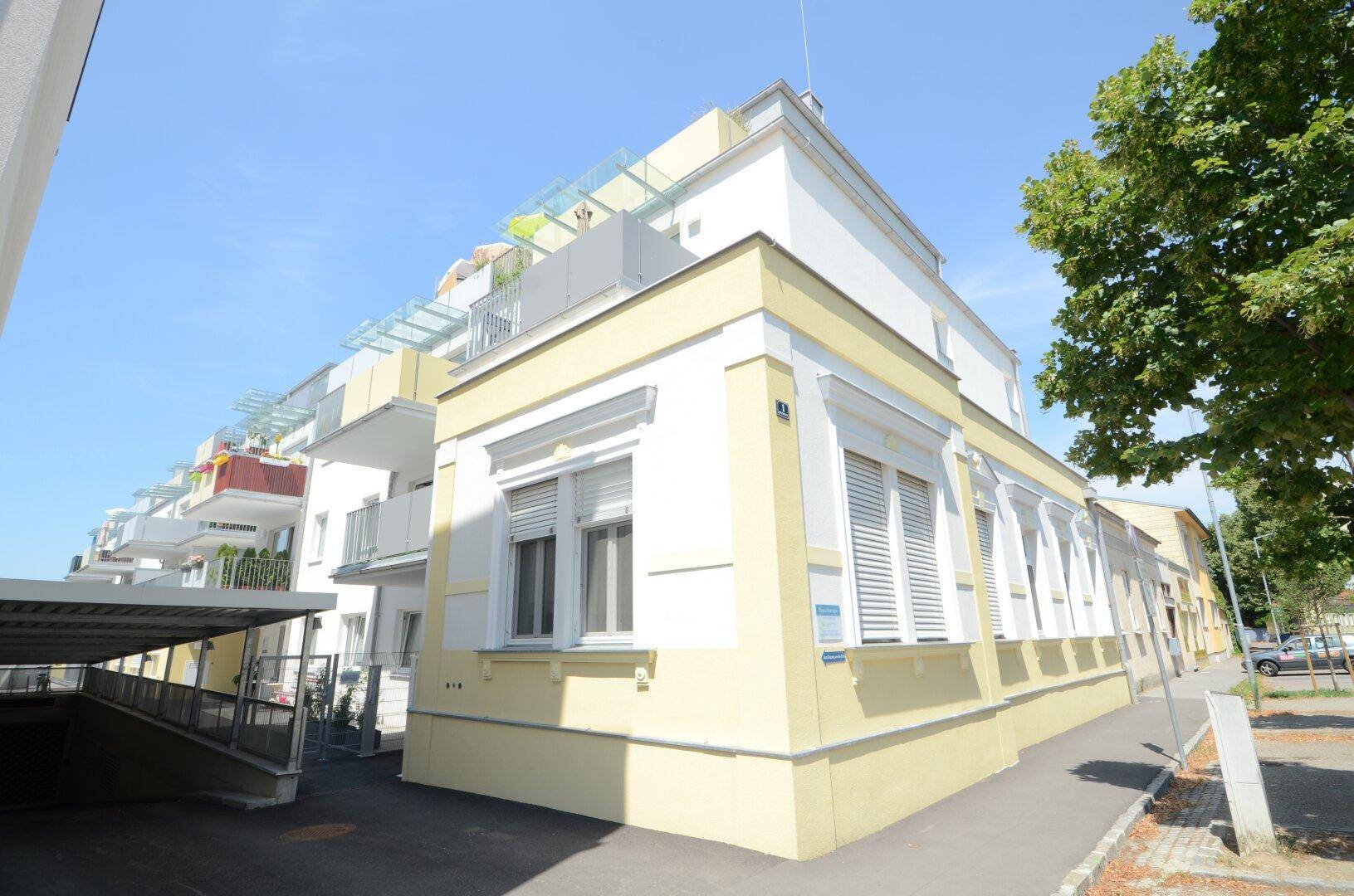 Gebäudeecke mit heller Fassade, Rollläden und der Einfahrt zu einer Tiefgarage an einem sonnigen Tag.