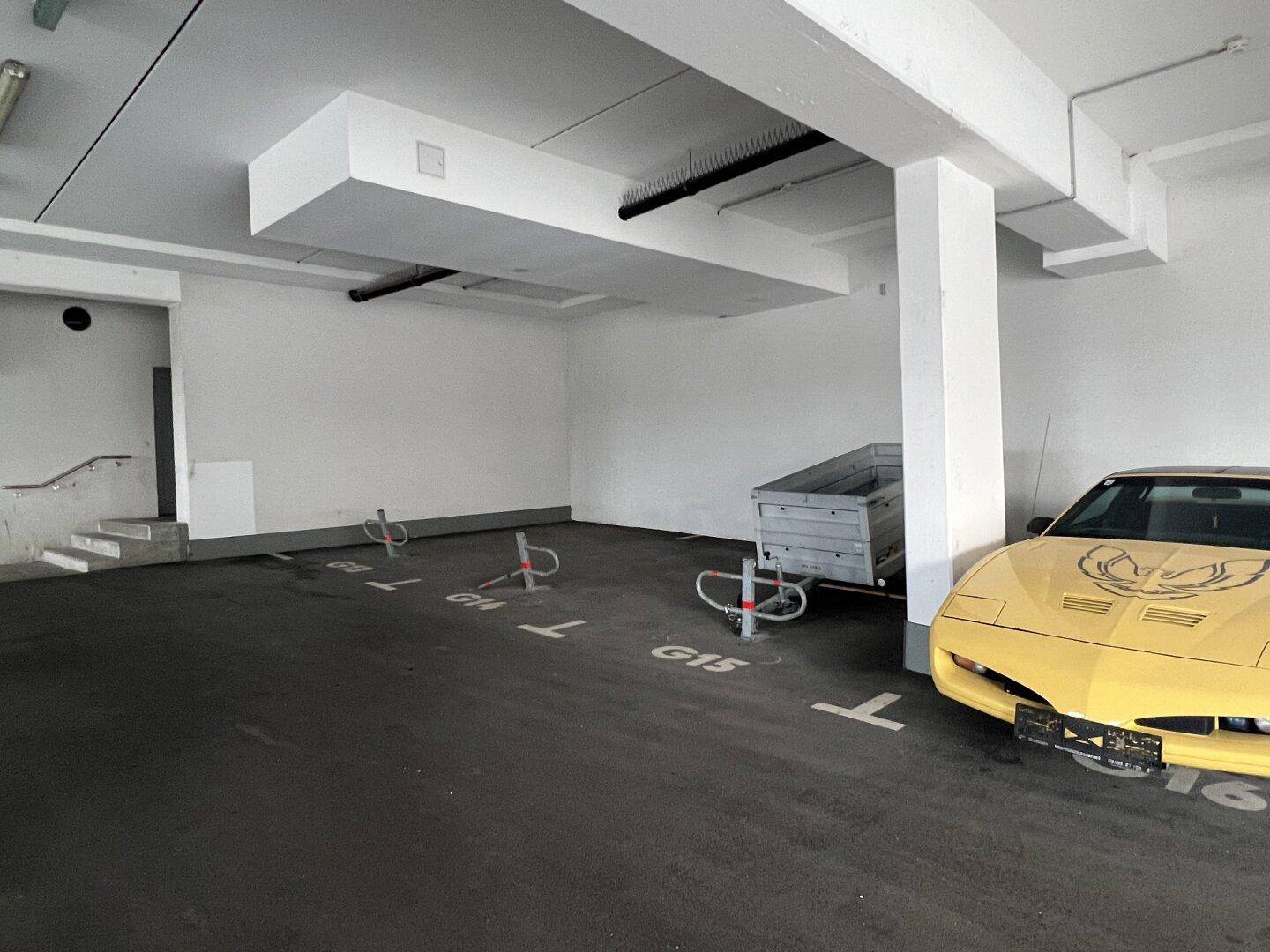 Tiefgarage mit markierten Stellplätzen, einem gelben Sportwagen und einem Anhänger, inklusive Treppenzugang.