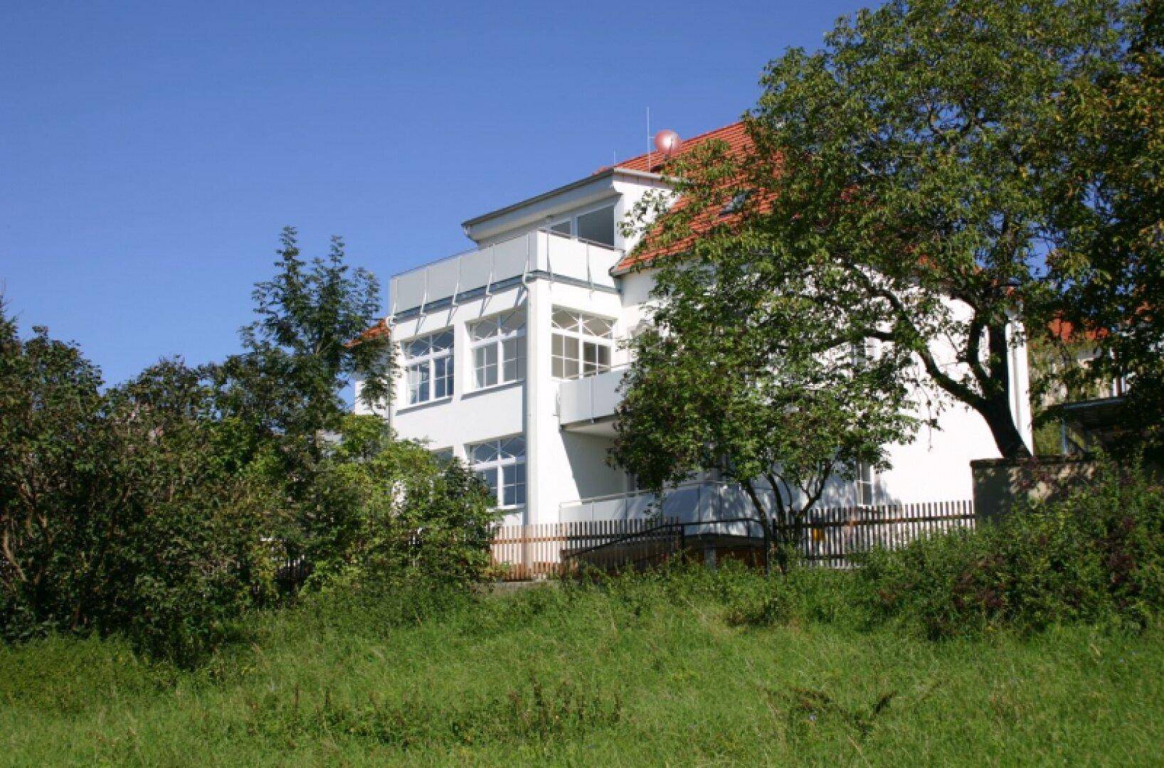 Weißes Gebäude mit Balkonen, umgeben von Bäumen und grüner Landschaft.
