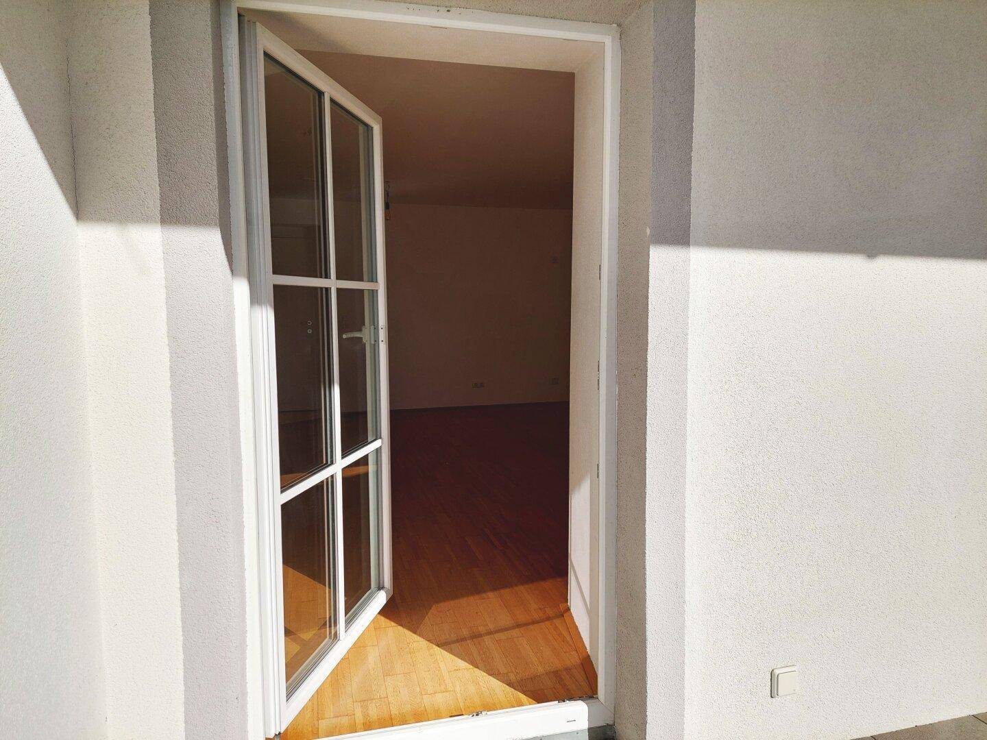 Blick vom Balkon durch eine offene Tür in einen leeren Raum mit Holzboden.