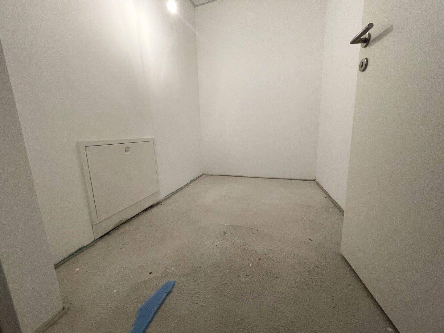 Kleiner, unfertiger Raum mit Betonboden und weißer Wand, ideal als Abstellraum oder Technikbereich.